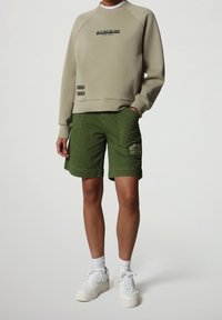 Sudadera beige con logo, combinada con pantalones cortos verdes que tienen bolsillos laterales y un parche. Se lleva con zapatillas blancas y calcetines.