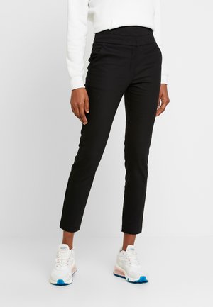 Vrouw draagt strakke zwarte broek tot de enkels, wit shirt met lange mouwen en witte sneakers met blauwe accenten, staand tegen een witte achtergrond.