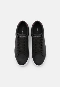 Sneakers in pelle nera con punte tonde, dettagli marroni sui talloni, suole in gomma bianca e lacci neri. Logo "Tommy Hilfiger" visibile all'interno.