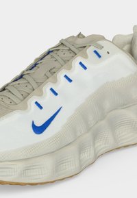 Λευκό και μπεζ αθλητικό παπούτσι Nike με μπλε λογότυπο Swoosh, μπλε ραφές, παχιά υφασμένη σόλα και ανοιχτό μπεζ κορδόνια.