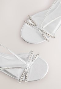 Sandales à talons aiguilles argentées avec une finition brillante, des lanières croisées ornées de strass transparents, et un design à bout ouvert.