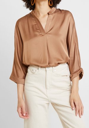 Mujer con una blusa marrón brillante de manga larga con puños enrollados y pantalones blancos de cintura alta, complementada con grandes pendientes de aro naranjas.
