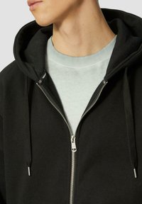 Schwarze Zip-Hoodie mit silbernem Reißverschluss, Kordelzügen und einem hellgrauen Rundhalsausschnitt darunter. Weicher Stoff und entspannte Passform.