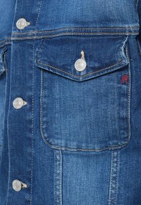 Jeansjacke mit Knopfleiste, silberner Hardware, Brusttaschen und rotem Stickdetail auf der Tasche. Klassische blaue Farbe und Textur.