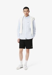 Lichtblauw overhemd met knopen, zwarte shorts, witte kabelgebreide sweater over de schouders, witte sneakers met crème accenten, met het merkelogo.