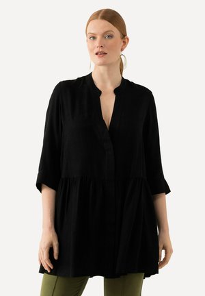 Donna con capelli rossi raccolti, che indossa grandi orecchini a cerchio, una tunica nera ampia con maniche 3/4 e pantaloni verde oliva, su uno sfondo bianco.
