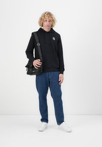 Hanorac negru din bumbac cu șnururi și un mic logo, asortat cu blugi albaștri și adidași albi. Modelul ține o geantă de umăr neagră.