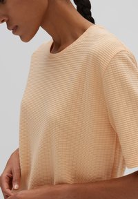 T-shirt di colore pesco con collo a giro e maniche corte. Il tessuto presenta un motivo a griglia in rilievo e si drappeggia delicatamente, mostrando la forma naturale.