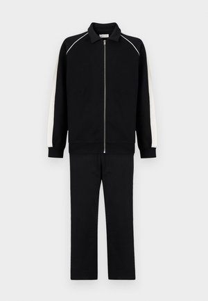 Ensemble de survêtement noir avec des rayures blanches sur les côtés, veste zippée intégrale avec col, et pantalon assorti à jambes droites sur un fond uni.