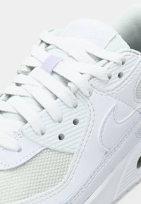 Zapatilla blanca con malla texturizada y cuero liso. Presenta cordones redondos blancos, un acento azul claro y una unidad de aire visible en la suela.