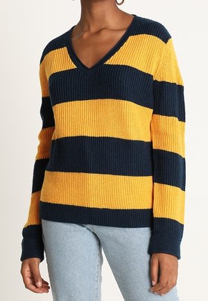 Pullover mit V-Ausschnitt und langen Ärmeln, mit breiten horizontalen Streifen in Marineblau und Senfgelb, getragen zu hellblauen Jeans.