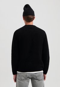 Schwarzer strukturierten Pullover mit langen Ärmeln und einem runden Ausschnitt, über einem weißen Hemd getragen. Graue Jeans und eine schwarze Mütze vervollständigen den Look.