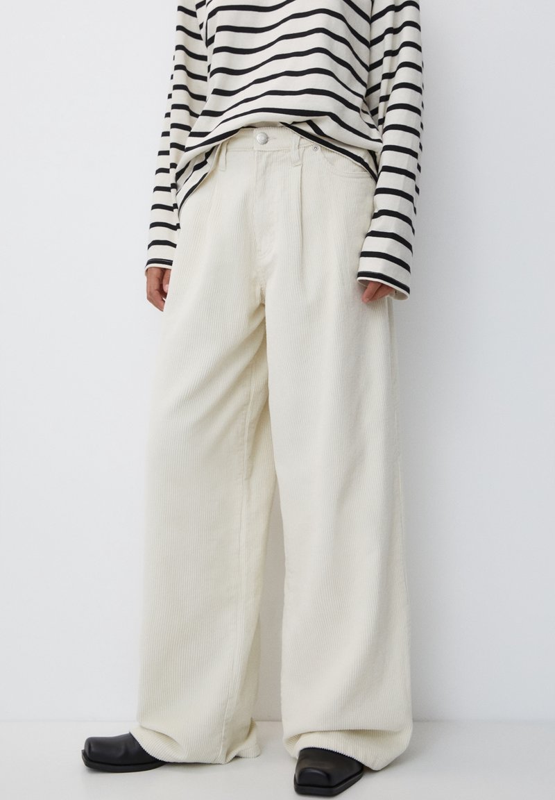 Personne portant un pantalon en velours côtelé crème à jambes larges, des chaussures noires à bout carré et un haut à manches longues rayé noir et blanc sur fond blanc.