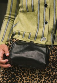 Bolso cruzado de cuero negro con textura suave y un sutil logo en relieve. Correa ajustable y diseño simple y estructurado.