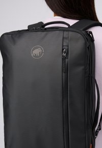 Svart rektangulär ryggsäck med framficka med dragkedja och mammutlogotyp, bärs av en person med långt mörkt hår och en ljusrosa skjorta.