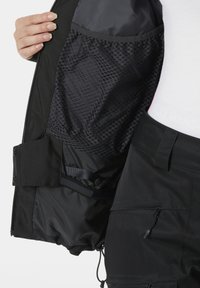Vnitřek černé bundy má prodyšnou síťovinovou podšívku a hladkou látku, na vnějšku jsou viditelné zipy a detaily s knoflíky.