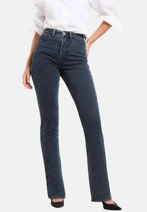 Mujer vestida con vaqueros de pierna recta en un oscuro azul de tiro alto, camisa blanca con las mangas remangadas y zapatos negros de punta, de pie con una mano en la cadera.