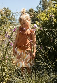 Bruine mouwloze top met kantafwerking en een witte rok met abstracte gele en bruine bladranden, opgesteld tussen hoge groene gras en bloemen.