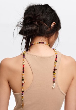 Kobieta o ciemnych włosach związanych w niski kok, ubrana w beżowy top na ramiączkach i kolorowy naszyjnik z koralików, spływający po jej plecach.