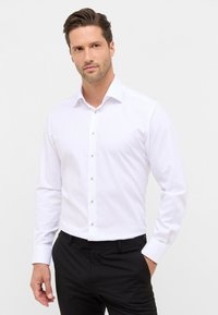 Man met een lichte huid en bruin haar, gekleed in een witte overhemd met lange mouwen en zwarte broek, met één hand in zijn zak.