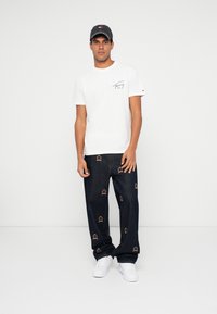 Hvit t-skjorte med grafisk design, mørkeblå jeans med brodert kronemønster, og hvite joggesko. Svart caps med logoelementer.