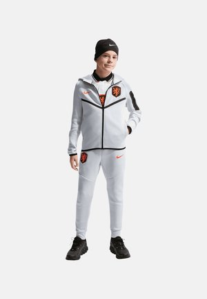 Ung person, der står, iført hvidt sportstøj med sorte og orange logoer, sort hue og sorte sportssko på en ensfarvet baggrund.
