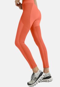Hoch taillierte orangefarbene Leggings mit strukturierten Einsätzen, figurbetontem Design und kontrastierenden Nähten, kombiniert mit weißen und schwarzen Sportschuhen.