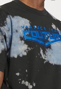 Schwarzes Batik-T-Shirt mit blauem "Versace Jeans Couture"-Logo. Kurze Ärmel, Rundhalsausschnitt, mit einer strukturierten Oberfläche und kontrastierenden Mustern.
