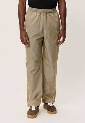 Beige træningsbukser lavet af let materiale, med elastisk talje, sidesømme og hvide kantdetaljer. Stylet med brune sneakers.