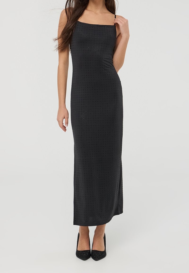 Femme portant une robe maxi noire moulante sans manches avec de petits ornements carrés et des chaussures noires à talons hauts, debout droite.