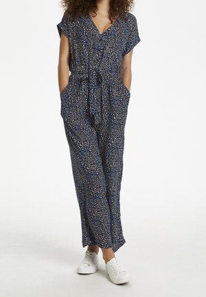 Kvinde iført en mørkeblå blomstret jumpsuit med korte ærmer og hvide sneakers, stående med hænderne i lommerne mod en hvid baggrund.