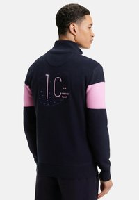 Pull en maille bleu marine avec un col montant, rayures roses aux épaules et un dos texturé présente un grand "10" et l'impression "French Flair".