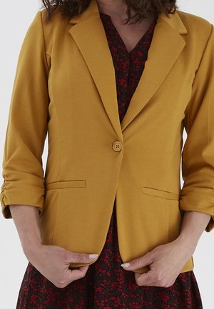 Woman wearing mustard yellow blazer with ruched sleeves over a black and red floral dress, hands adjusting the blazer front.