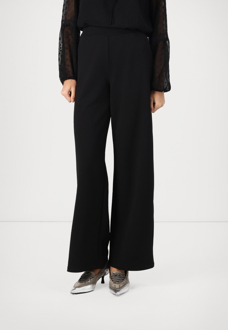 Femme portant un pantalon noir à jambes larges, une blouse noire transparente à manches longues et des escarpins métalliques argentés à bout pointu, debout sur un fond blanc.