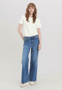 Kevyt valkoinen t-paita yhdistettynä leveälahkeisiin sinisiin farkkuihin. Farkuissa on perinteinen viiden taskun muotoilu ja ne ovat hieman haalistuneet.