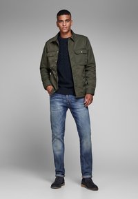 Groene utility-jack met borstzakken, marineblauwe kabelgebreide trui, vervaagde blauwe jeans en donkerbruine suède enkellaarzen. Soepele texturen, casual pasvorm.