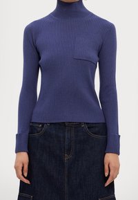 Geribbelde coltrui in diep paars, met lange mouwen en een borstzak, gecombineerd met een donkere denim rok. Zachte textuur, getailleerd design.