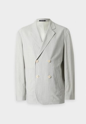 Jeune homme portant un blazer croisé gris clair, une chemise blanche, un pantalon noir à jambes larges et des chaussures noires, debout devant un fond uni.