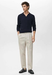 Young man standing with hands in pockets wearing a navy ribbed sweater, beige trousers, and black loafers against a plain light background.