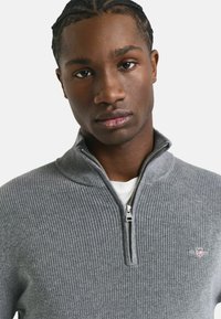 Junger Mann mit geflochtenem Haar, der einen grauen, gerippten Reißverschluss-Pullover über einem weißen Shirt vor einem schlichten Hintergrund trägt.