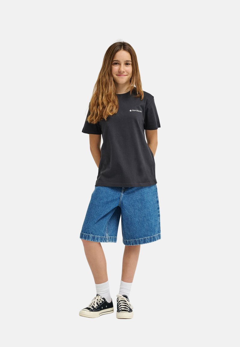 Joven de pie con las manos detrás de la espalda, lleva una camiseta negra de New Haven, pantalones cortos de mezclilla hasta la rodilla, calcetines blancos y zapatillas negras.