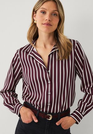 Vrouw met blond haar draagt een bordeauxrode en witte verticaal gestreepte blouse met knopen, een zwarte broek, gouden ronde oorbellen en een bordeauxrode riem.