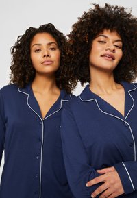 Mörkblå pyjamasöverdelar med vita detaljer, knäppning fram och lång ärm; en har en bröstficka. Texturen ser mjuk och bekväm ut.