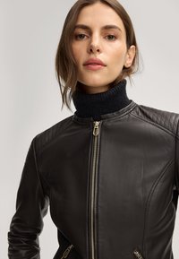 Schwarze Lederjacke mit gesteppten Schultern, taillierter Passform, hohem Kragen und goldenen Reißverschlussdetails. Geschmeidige Textur und elegante Silhouette.