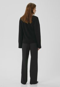 Sieviete ar gariem brūniem matiem, tērpusies melnā džemperī un platām melnām biksēm, stāvot ar muguru pret vienkāršu fona virsmu.