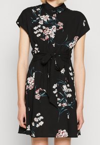 Schwarzes Kleid mit kurzen Kappenärmeln, Blumenmuster in Weiß und Pink, Knopfleiste vorne, taillierter Schnitt mit Bindegürtel, fließender Stoff, knielange Länge.