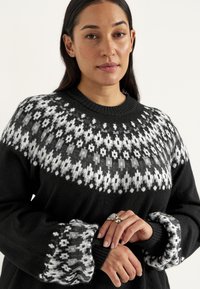 Sort sweater med hvidt geometrisk mønster omkring halsudskæringen og manchetterne. Har en afslappet pasform, ribbet krave og mønstret ærmeender.