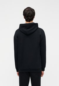 Μαύρο βαμβακερό φούτερ με χαλαρή εφαρμογή, με μεγάλη κουκούλα, ριμπ μανσέτες και τελείωμα. Η πίσω όψη δείχνει λεία υφή χωρίς σχέδια.