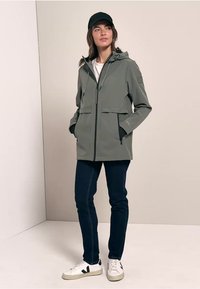 Veste légère de couleur olive avec capuche, deux poches avant et zippers ; assortie à un jean foncé et des baskets blanches.