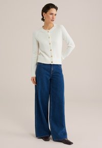 Vitt kardigan med rund hals, sju-knappars fram, ribbade manschetter, kombinerad med vida blå denimjeans och mörka skor. Slät textur.